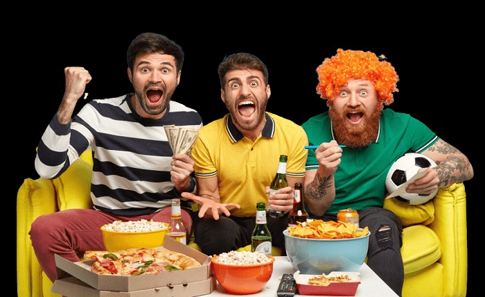 Drie mannen kijken enthousiast naar een sportwedstrijd via een IPTV-service met TiviMate Premium. Ze zitten op een gele bank met drankjes en snacks. Eén van hen houdt geld vast, een ander een voetbal, en de derde draagt een grappige oranje pruik. Ze zien er blij uit na hun abonnement via de website tivimate premium kopen